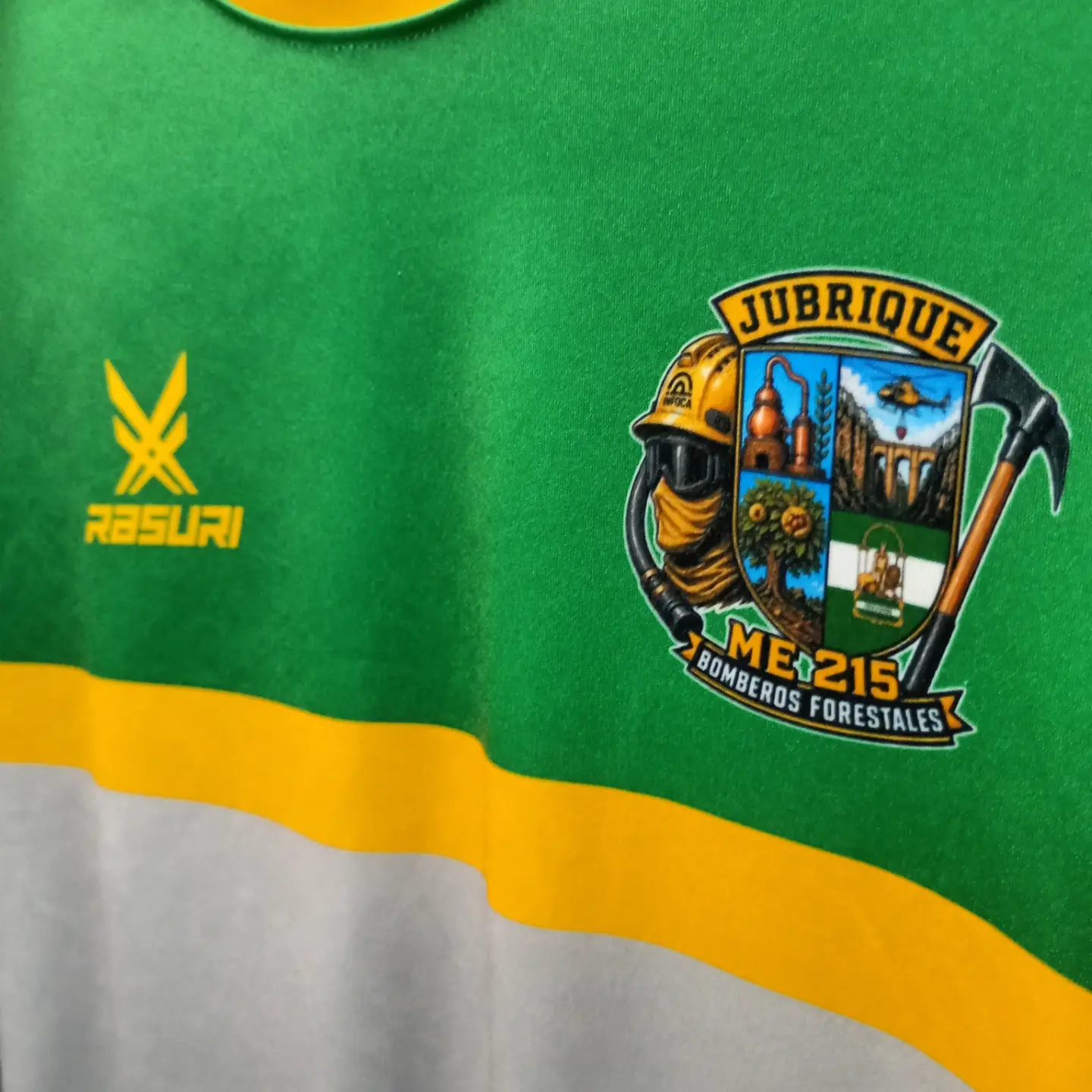 Detalle de una camiseta verde y amarilla con el escudo de los Bomberos Forestales de Jubrique y el logotipo Rasuri